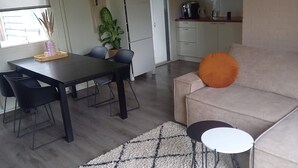 Une cuisine moderne avec des meubles blancs, un coin repas avec une table et des chaises noires, et un salon avec un canapé beige et un coussin orange.