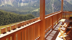 Ein Holzbalkon mit Tisch und Stühlen, der einen Blick auf eine gebirgige Landschaft mit einem bewaldeten Tal und schneebedeckten Gipfeln bietet.