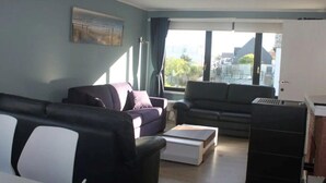 A modern living room with two sofas, a coffee table, and a kitchen area with a refrigerator. - Cozy Family Duplex on the Belgian Coast (Middelkerke)
