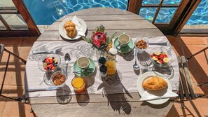 Une table de petit-déjeuner avec des croissants, des fruits et des boissons, installée au bord d’une piscine avec vue sur l’eau.