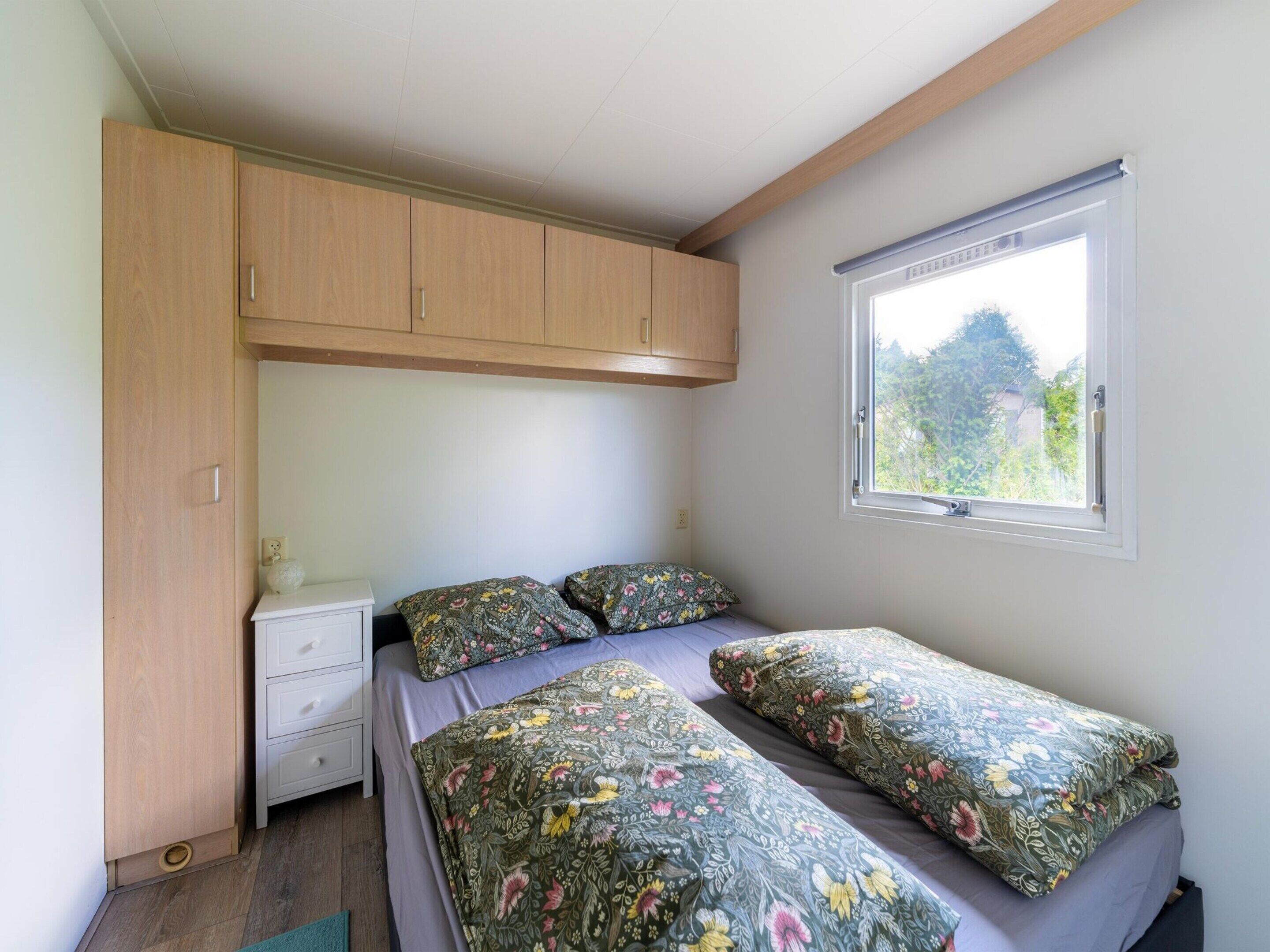 A small, well-lit bedroom with a single bed, a nightstand, a wooden wardrobe, and a window providing a view of greenery outside.