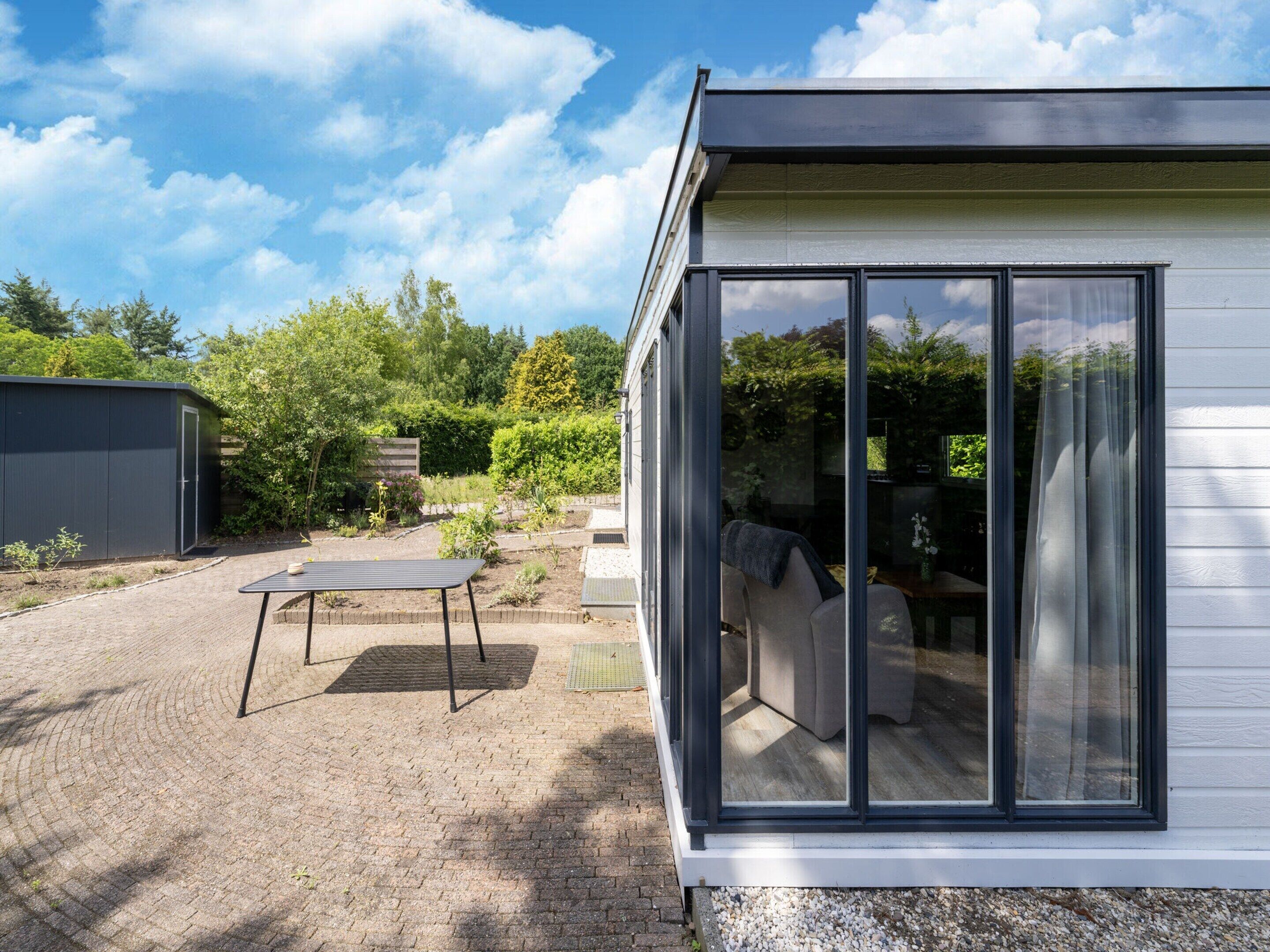 A modern house with a large glass window, a patio with a table and chairs, and a garden area.