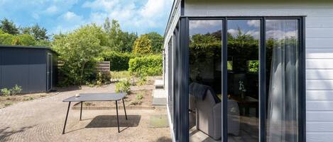 A modern house with a large glass window, a patio with a table and chairs, and a garden area.