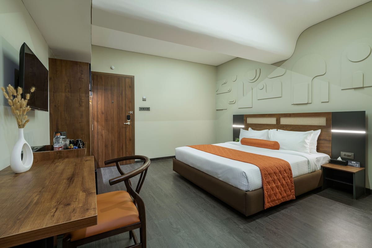A hotel room with a wooden desk, a bed with an orange blanket, a nightstand, and a wall-mounted TV.