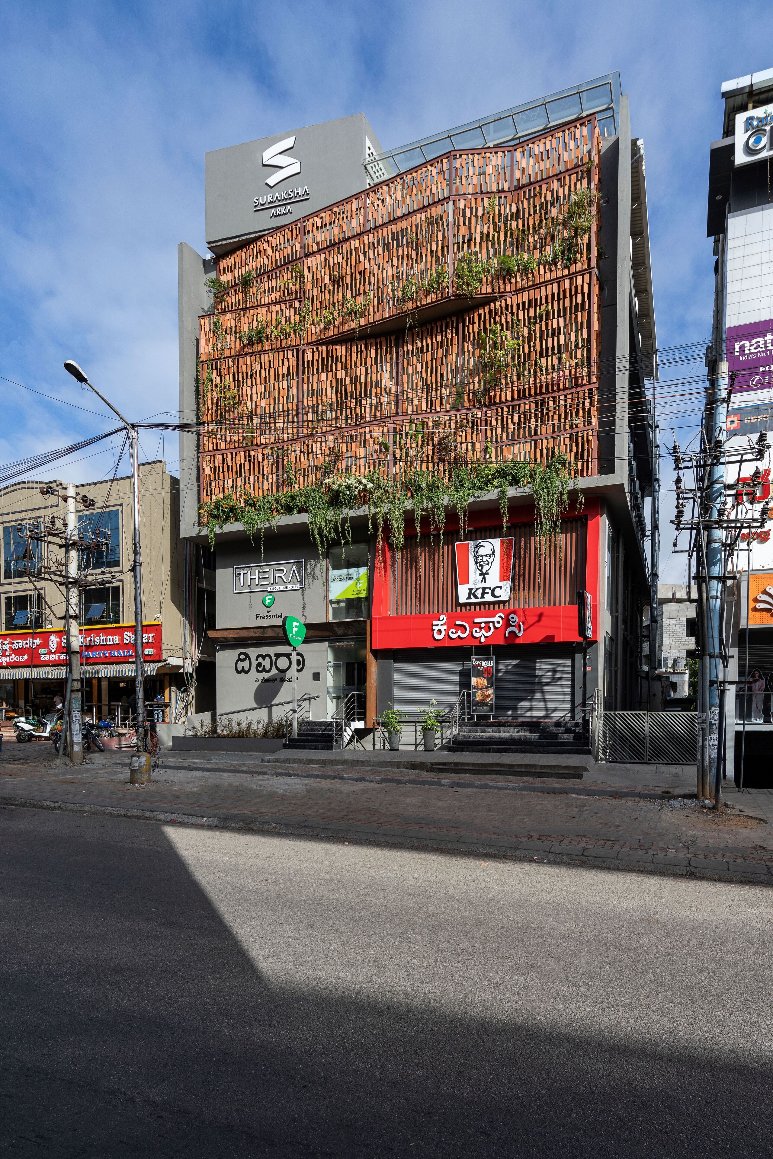 A building with a KFC restaurant on the ground floor, a sign for Suraksha Arka, and a street with parked motorcycles.