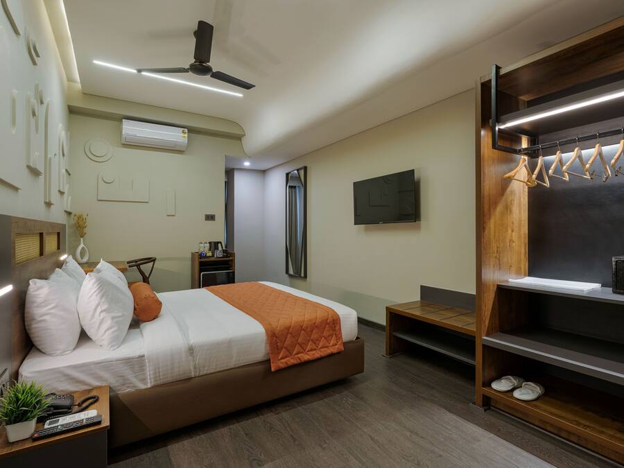 Une chambre d’hôtel moderne équipée d’un lit, d’une télévision, d’un ventilateur de plafond, d’une étagère en bois avec des cintres et d’une petite table sur laquelle se trouvent une plante et des télécommandes.