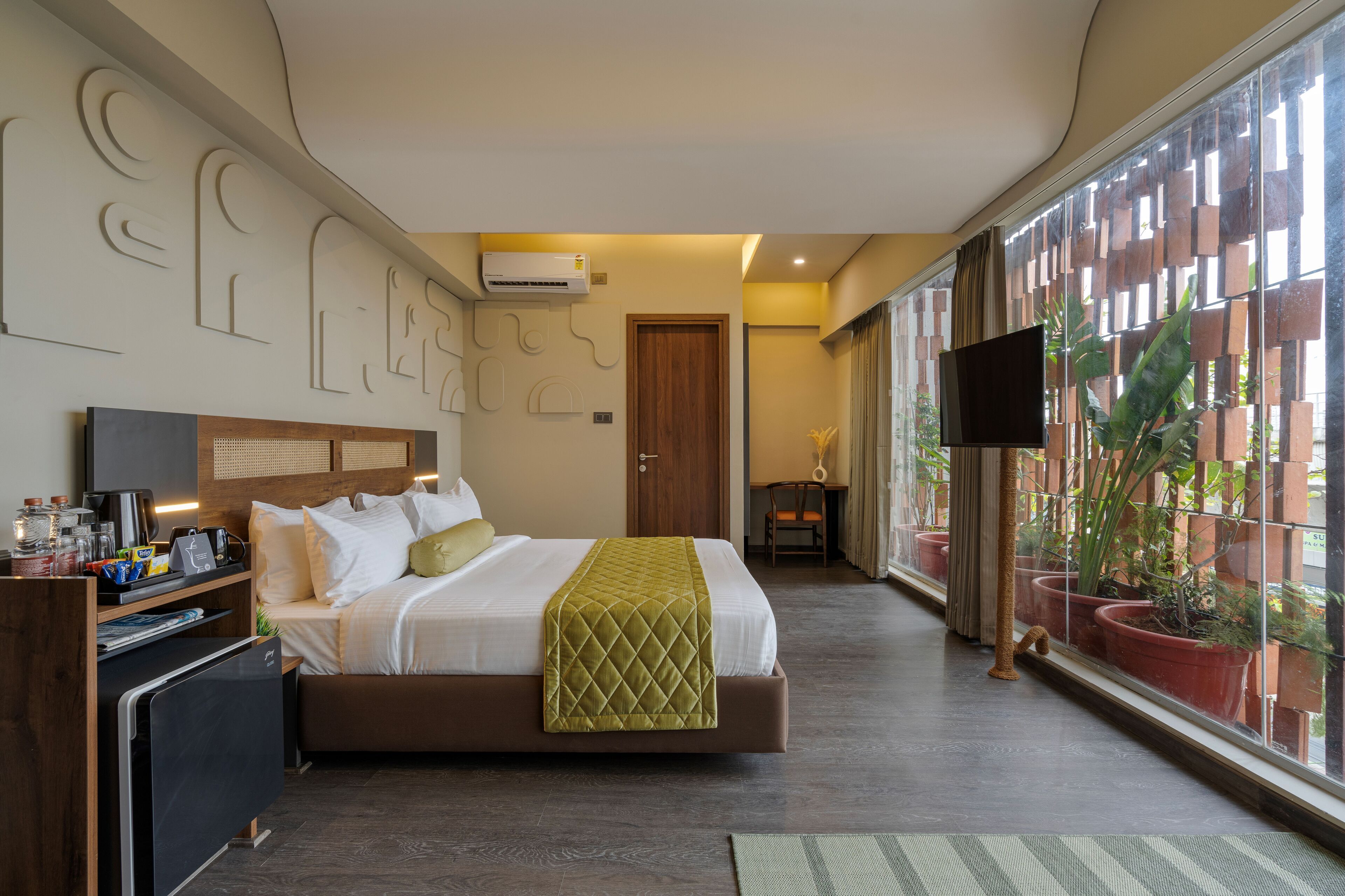 A hotel room with a bed, a desk, a TV, and a large window with plants.