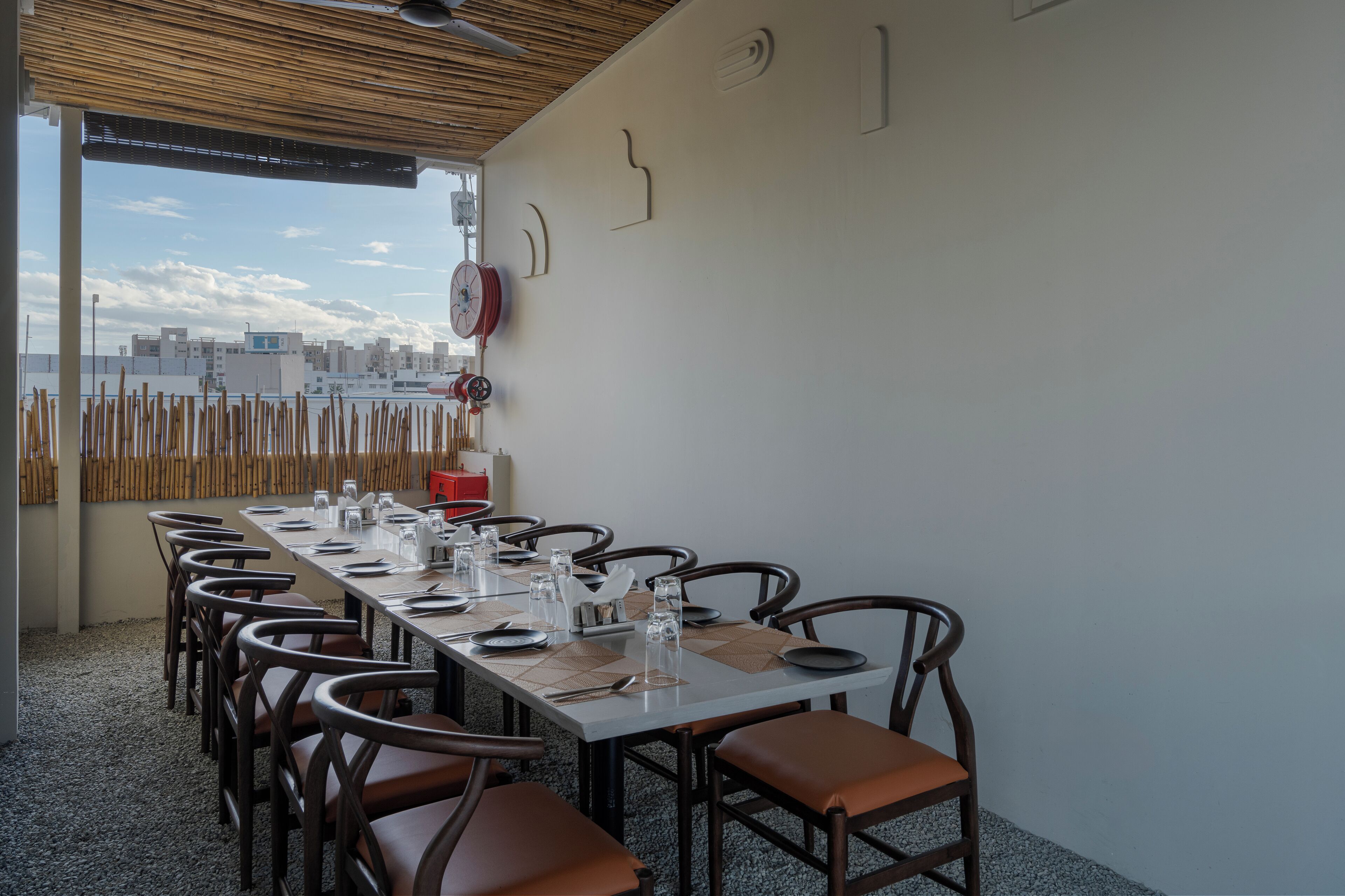 A dining area with a long table set for a meal, surrounded by chairs, with a cityscape view through an open wall.