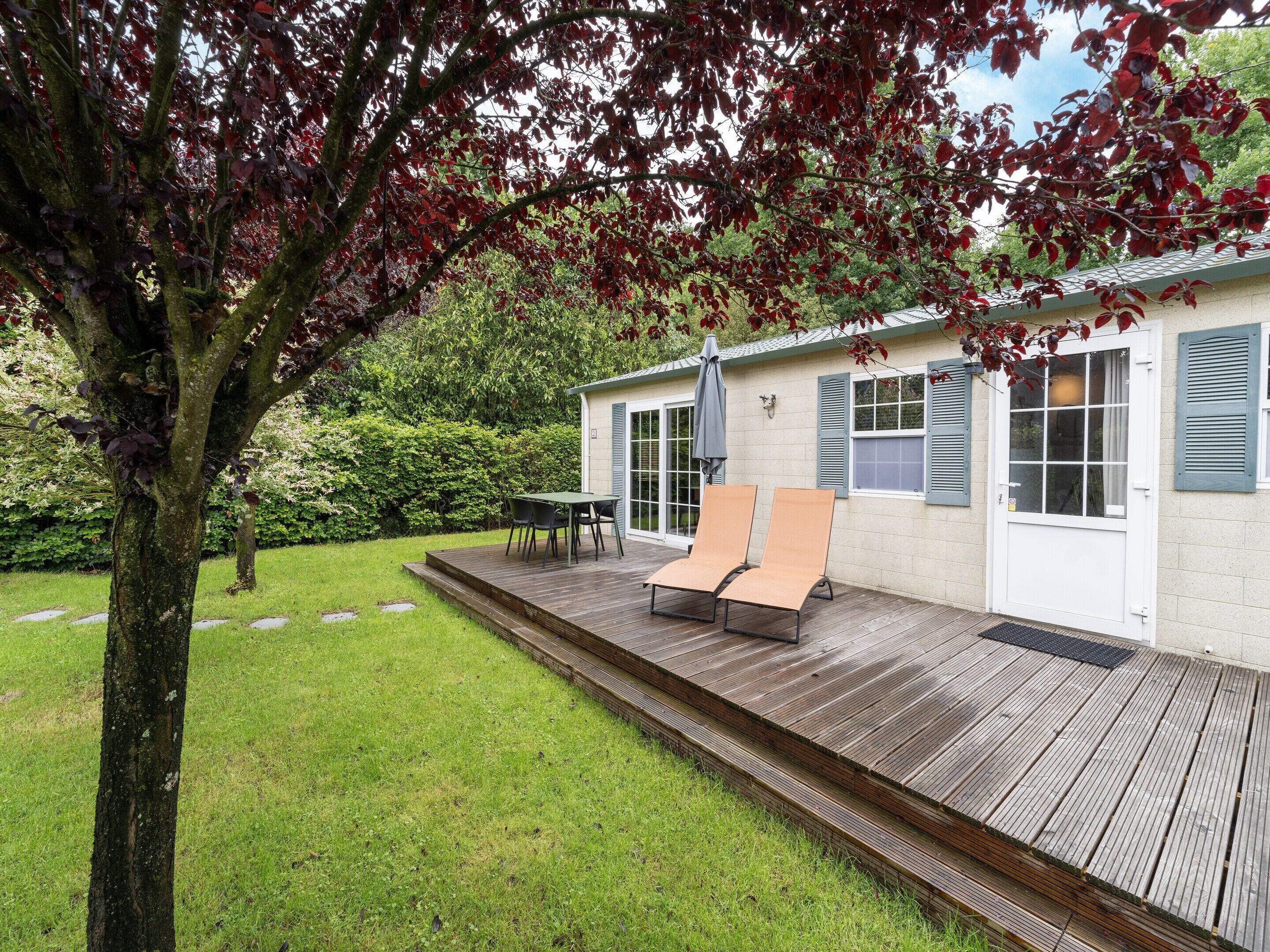 Eine Holzterrasse mit zwei Liegestühlen und einem Tisch, ein kleiner Garten und ein Gebäude mit blauen Fensterläden.