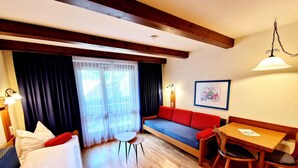 Una habitación acogedora con piso de madera, un sofá con cojines rojos, una mesita con sillas y una ventana con cortinas translúcidas.
