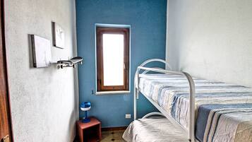 A room with a bunk bed, a small window, a door, and a wall-mounted shelf with a lamp.
