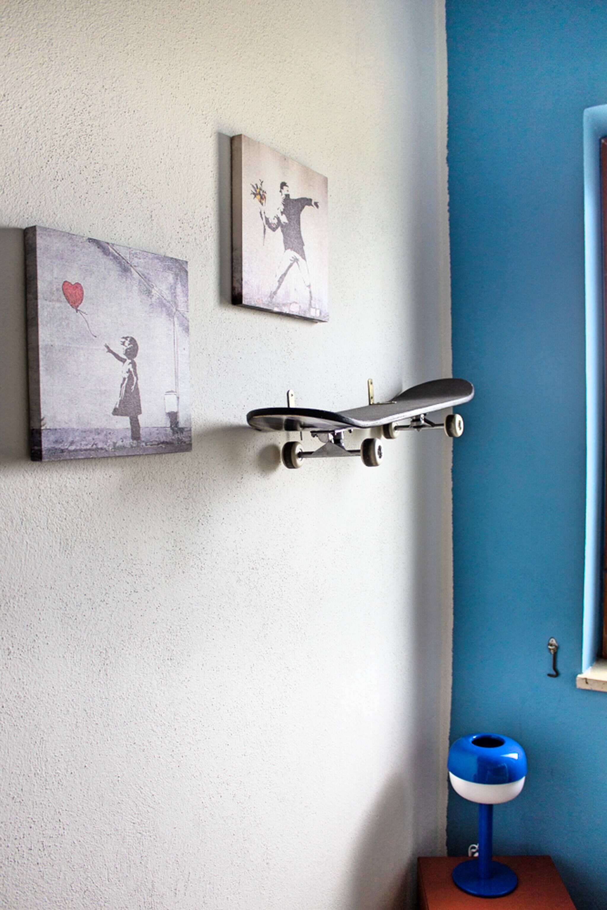 Una pared con dos cuadros enmarcados, un monopatín fijado a la pared y un objeto azul y blanco sobre una mesita.