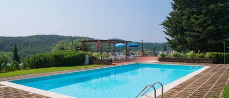A rectangular swimming pool with a tiled deck, surrounded by greenery and a pergola with umbrellas.