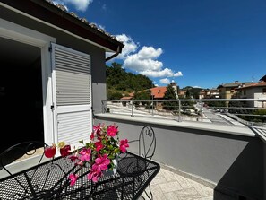En balkon med et bord og stole, en potteplante med lyserøde blomster og udsigt til et boligområde med huse og træer.