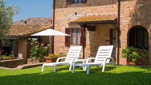 A stone building with a tiled roof, two white lounge chairs, and potted plants.