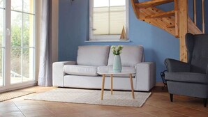Una sala de estar con un sofá gris, una mesita con un florero, un sillón gris, una escalera de madera y una ventana con persianas.