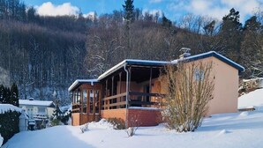 A chalet-style building with a wooden deck and a large window, surrounded by snow-covered ground and trees. - Ferienwohnung mit Terrasse in Blankenburg (Blankenburg)