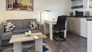 A modern living room with a grey sofa, a small white table, and a black chair. There is a painting of a forest on the wall, a white cabinet with glass doors, and a microwave on the counter. - Ferienwohnung mit Terrasse in Blankenburg (Blankenburg)