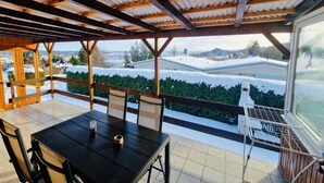 Un espace extérieur abrité, aménagé pour les repas, avec une table et des chaises, une vue sur des maisons enneigées et une structure en pergola en bois.