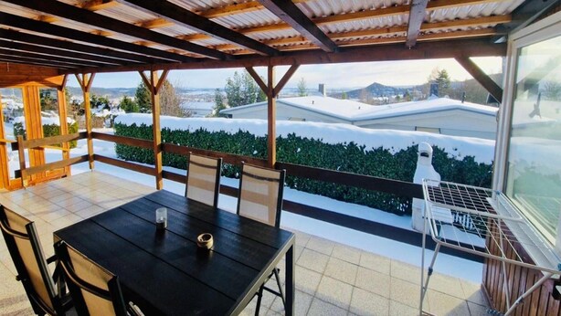 A covered outdoor dining area with a table and chairs, a view of snow-covered houses, and a wooden pergola structure. - Ferienwohnung mit Terrasse in Blankenburg (Blankenburg)