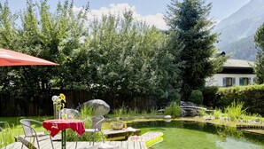 A wooden deck with a table and chairs, a small pond, and a house in the background. - Maximilian 1-2 People (Neukirchen am Grossvenediger)