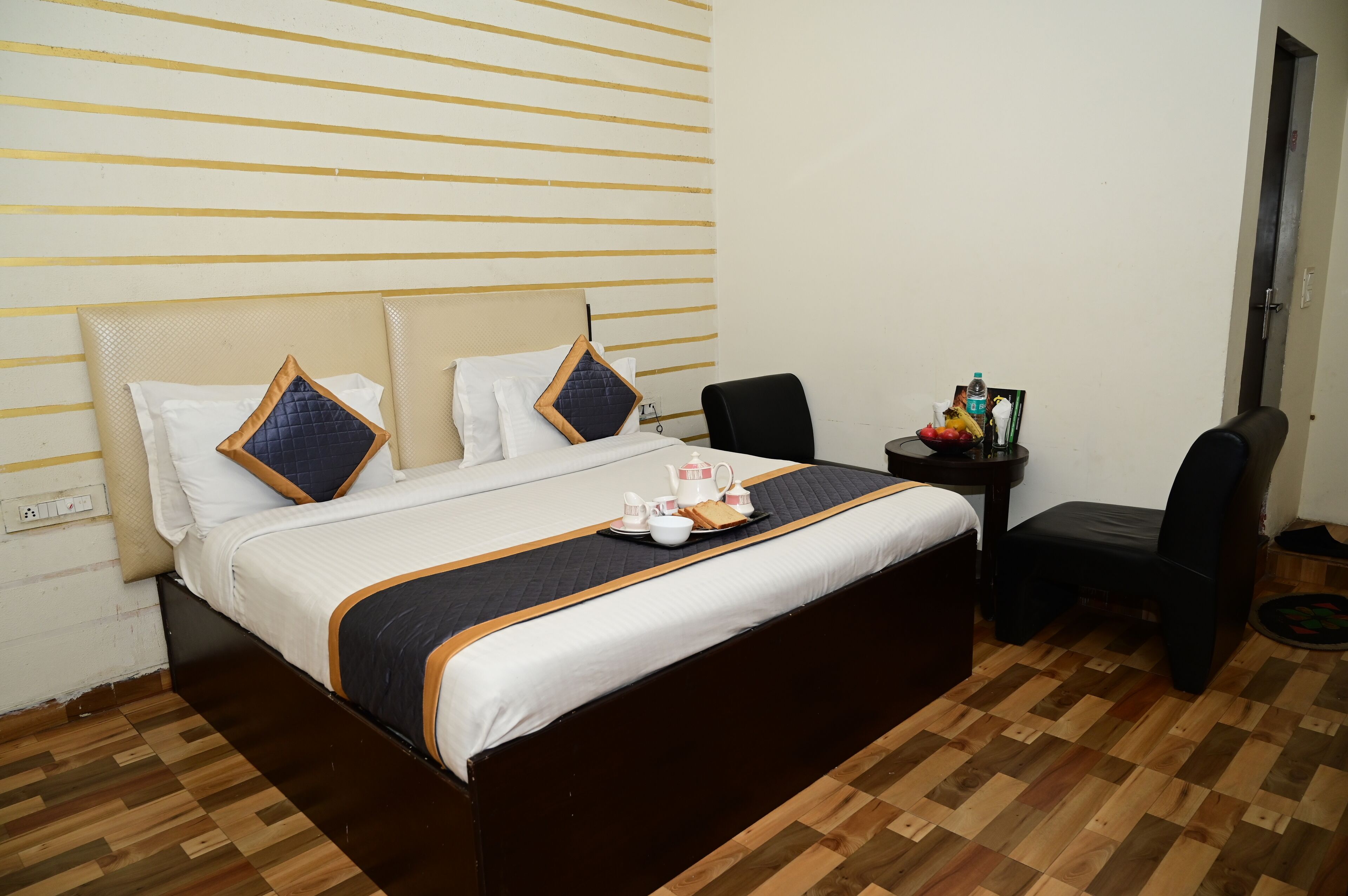 A neatly made bed with a tray of breakfast items, a chair, and a small table with a bottle and a picture frame.