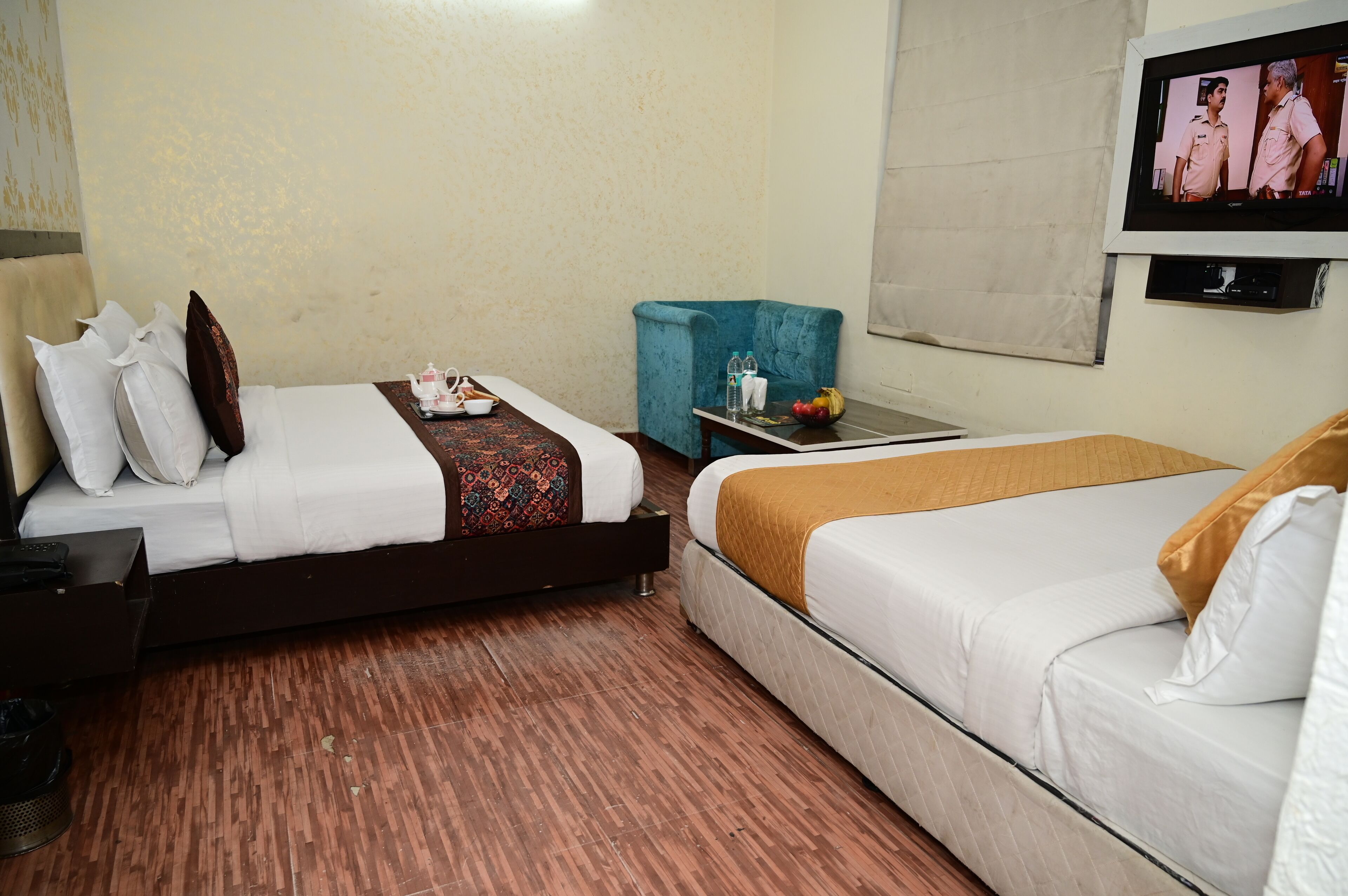 A hotel room with two beds, a TV, a small table with a fruit bowl, and a blue chair.