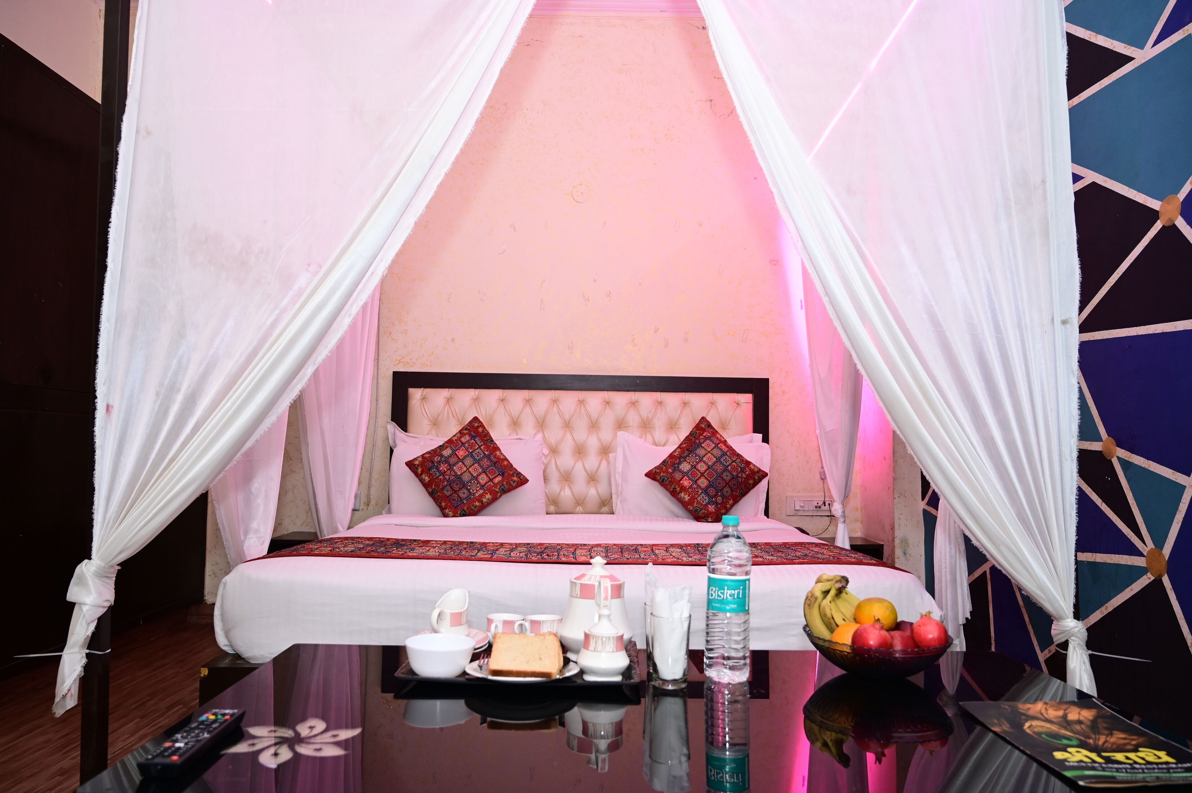 A hotel room with a bed, a table with breakfast items, a bottle of water, and a fruit bowl.