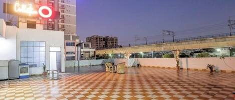 A rooftop terrace with a checkered floor, a covered walkway, and a building with a large illuminated sign.