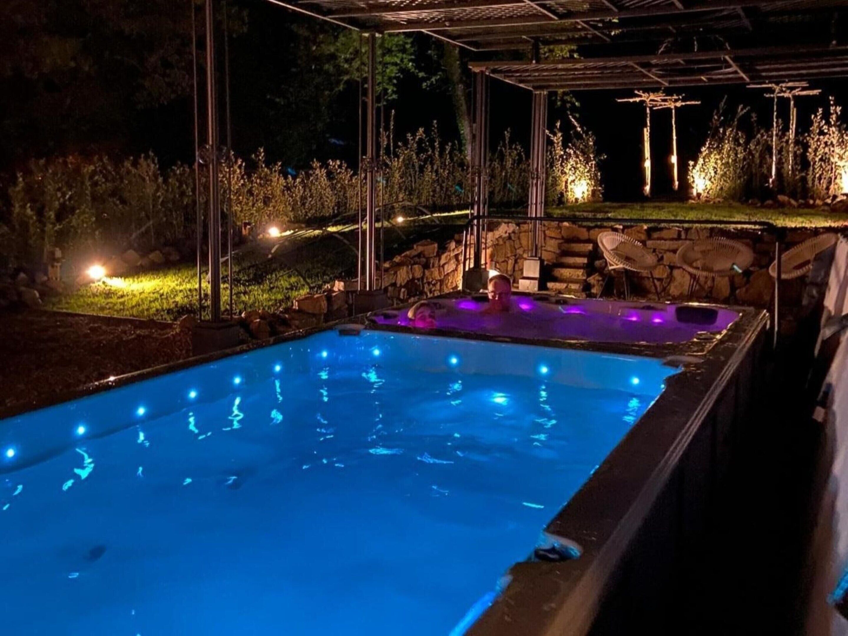 Ein Schwimmbecken mit Whirlpoolbereich, umgeben von Steinmauern und Pflanzen, unter einer Überdachung bei Nacht.