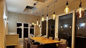 A modern dining area with a wooden table and wicker chairs, hanging light fixtures, and a view through large sliding glass doors. - Luxurious Villa With Heated Pool (Gesves)