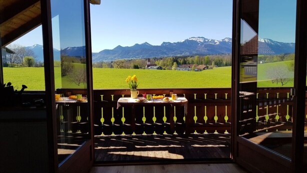 A room with a wooden floor, a table with a vase of flowers, and a view of mountains and a green field. - Mountain View Comfortable Holiday Residence (Rimsting)