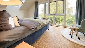 Un dormitorio con una cama grande, una mesita, una silla y una ventana con vistas a árboles.