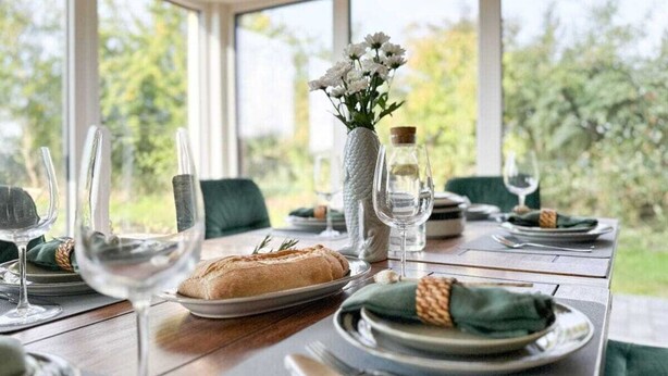 Una mesa de comedor dispuesta con platos, vasos y una hogaza de pan, con un jarrón de flores y una botella de agua.