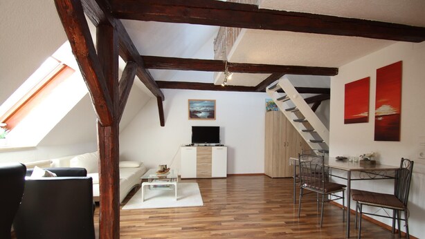 A loft-style living space with exposed wooden beams, a white sofa, a dining table with chairs, and artwork on the walls. - Behagliche Ferienwohnung in Elbingerode (Oberharz am Brocken)