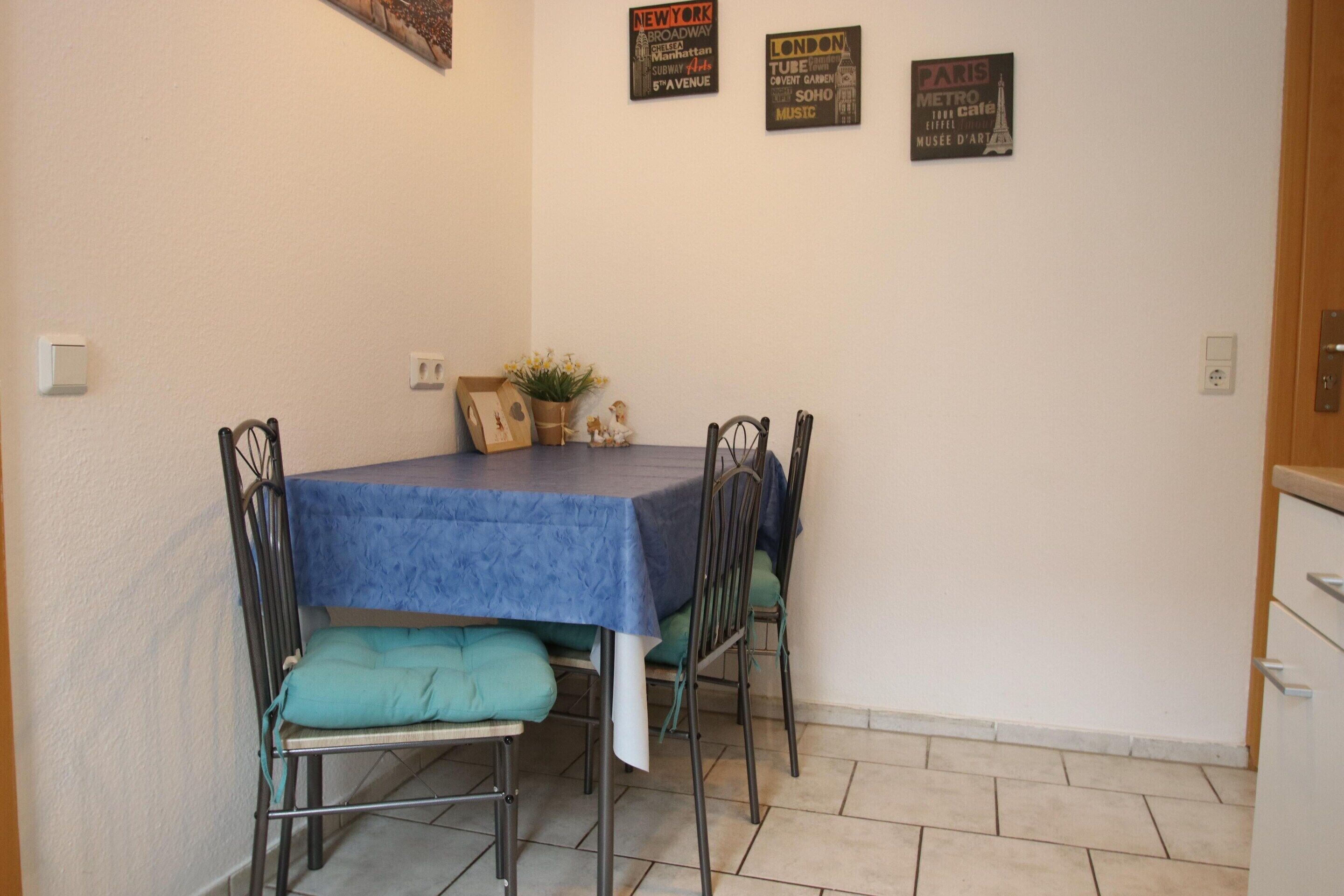 Ein Essbereich mit einem Tisch, der mit einer blauen Tischdecke bedeckt ist, vier Stühle mit grünen Kissen und Wanddekorationen, darunter Schilder für New York, London und Paris.