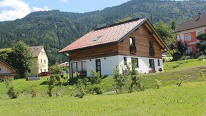 A two-story wooden house with a red-tiled roof, surrounded by a green landscape and a forested mountain in the background. - Huis 3landenpunt AT-SLO-IT. Holiday in Austria, Slovenia and Italy at once. (Arnoldstein)