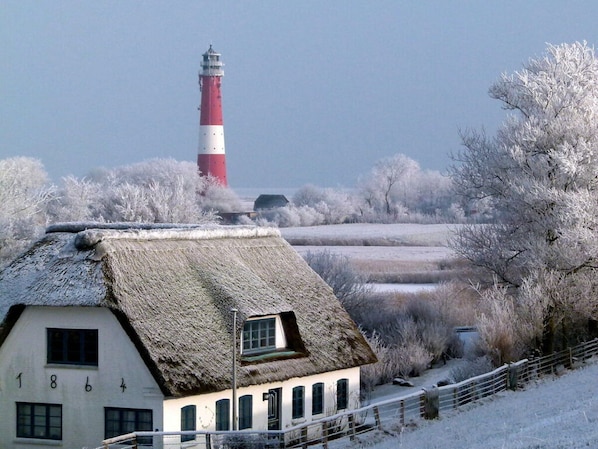 A thatched-roof house with a visible number '1864' on the side, a red and white lighthouse in the background, and frost-covered trees and fields. - Pellworm Ostersiel 1 (Pellworm)