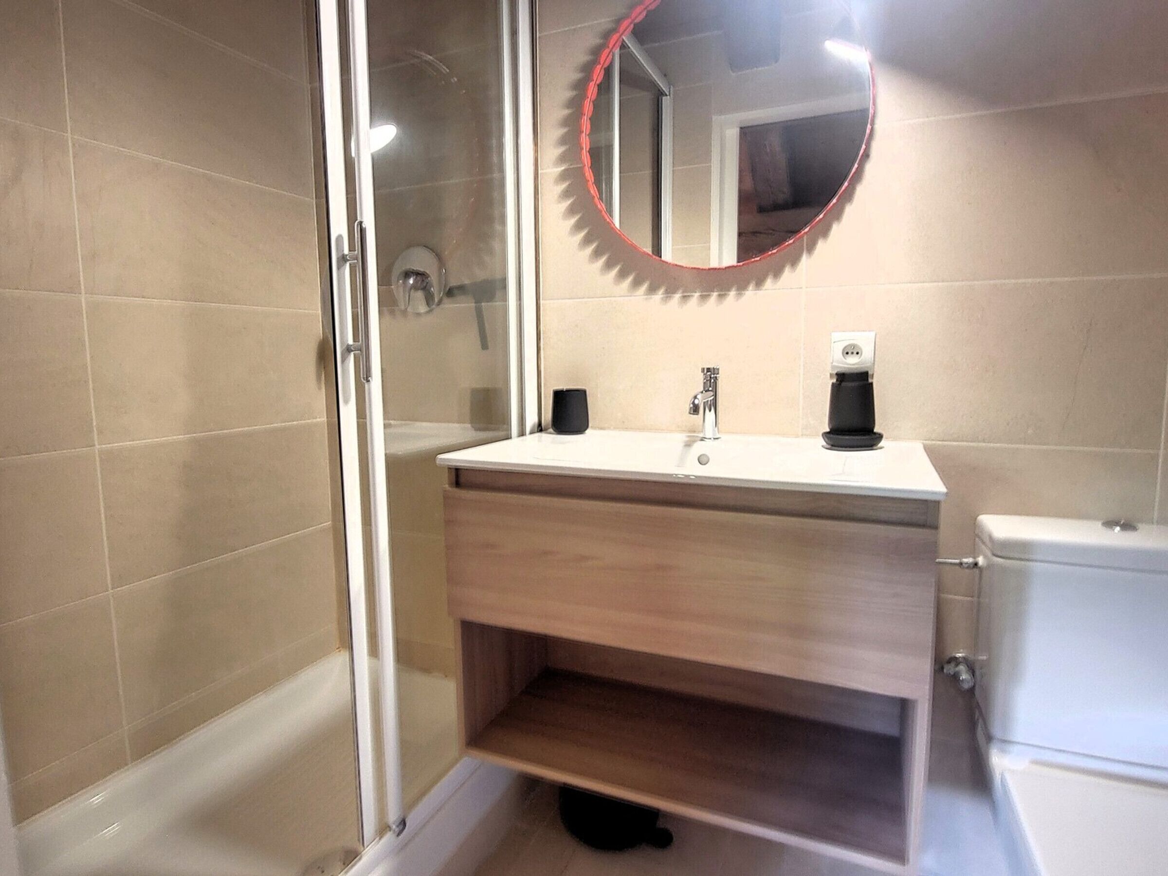 A modern bathroom with a glass shower enclosure, a white sink with a black faucet, and a round mirror with a red frame.