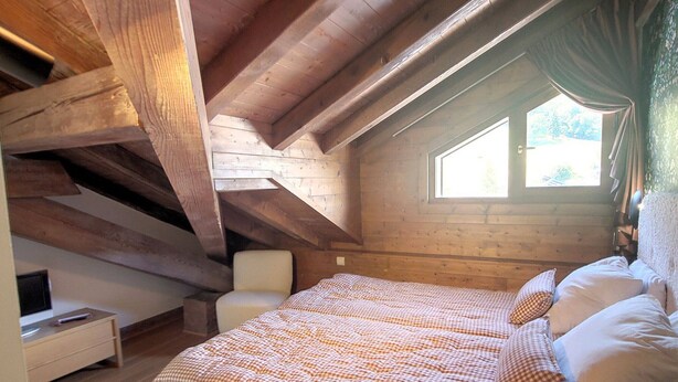 Een gezellig zolderkamertje met een geruit beddegoed, een witte stoel en een houten plafond.