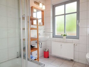 Une salle de bain avec une douche à parois de verre, une étagère en bois où sont rangés les articles de toilette, un lavabo blanc et une fenêtre avec vue.
