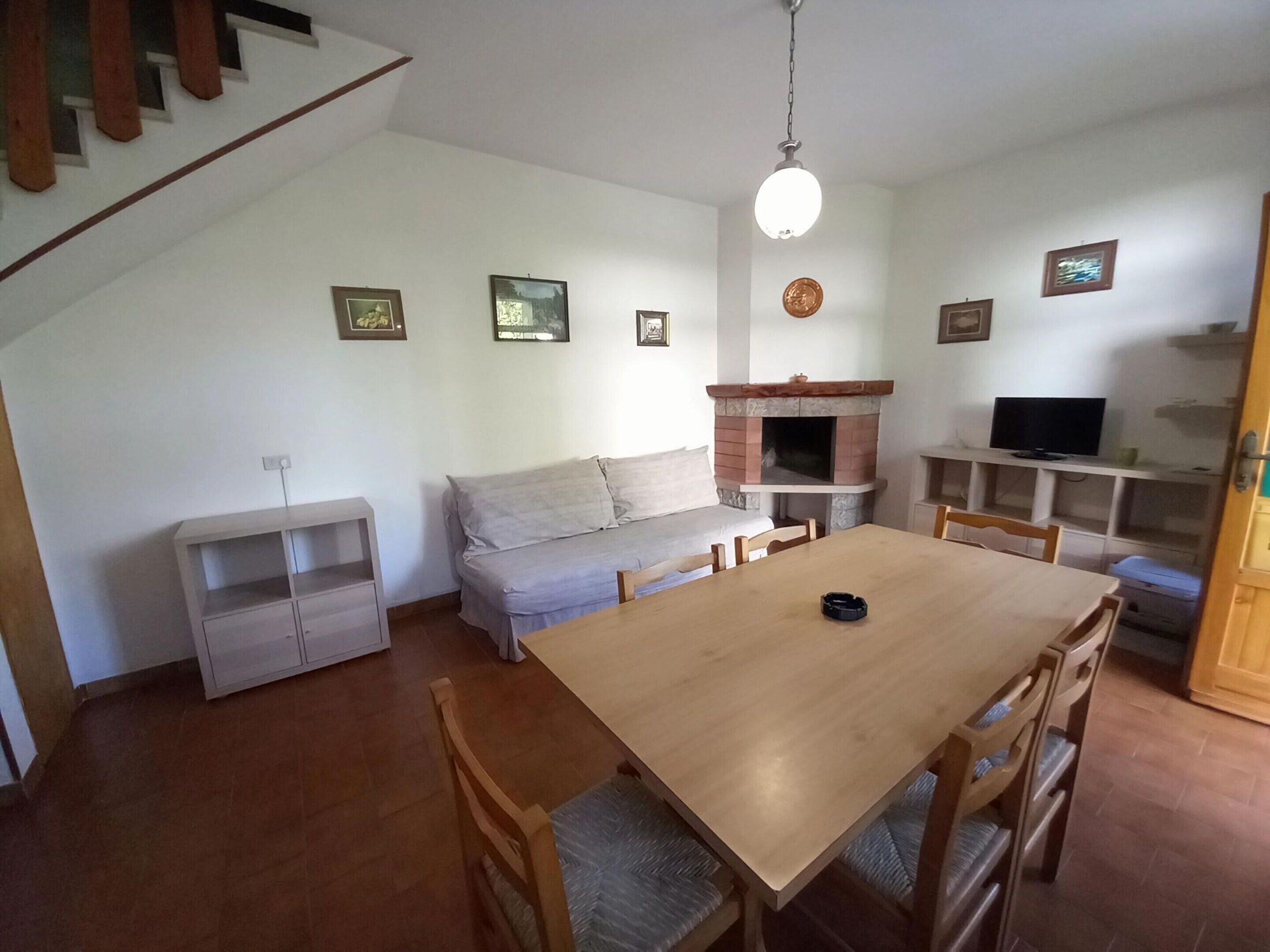 A dining area with a wooden table and chairs, a sofa, a fireplace, and a television on a shelf.