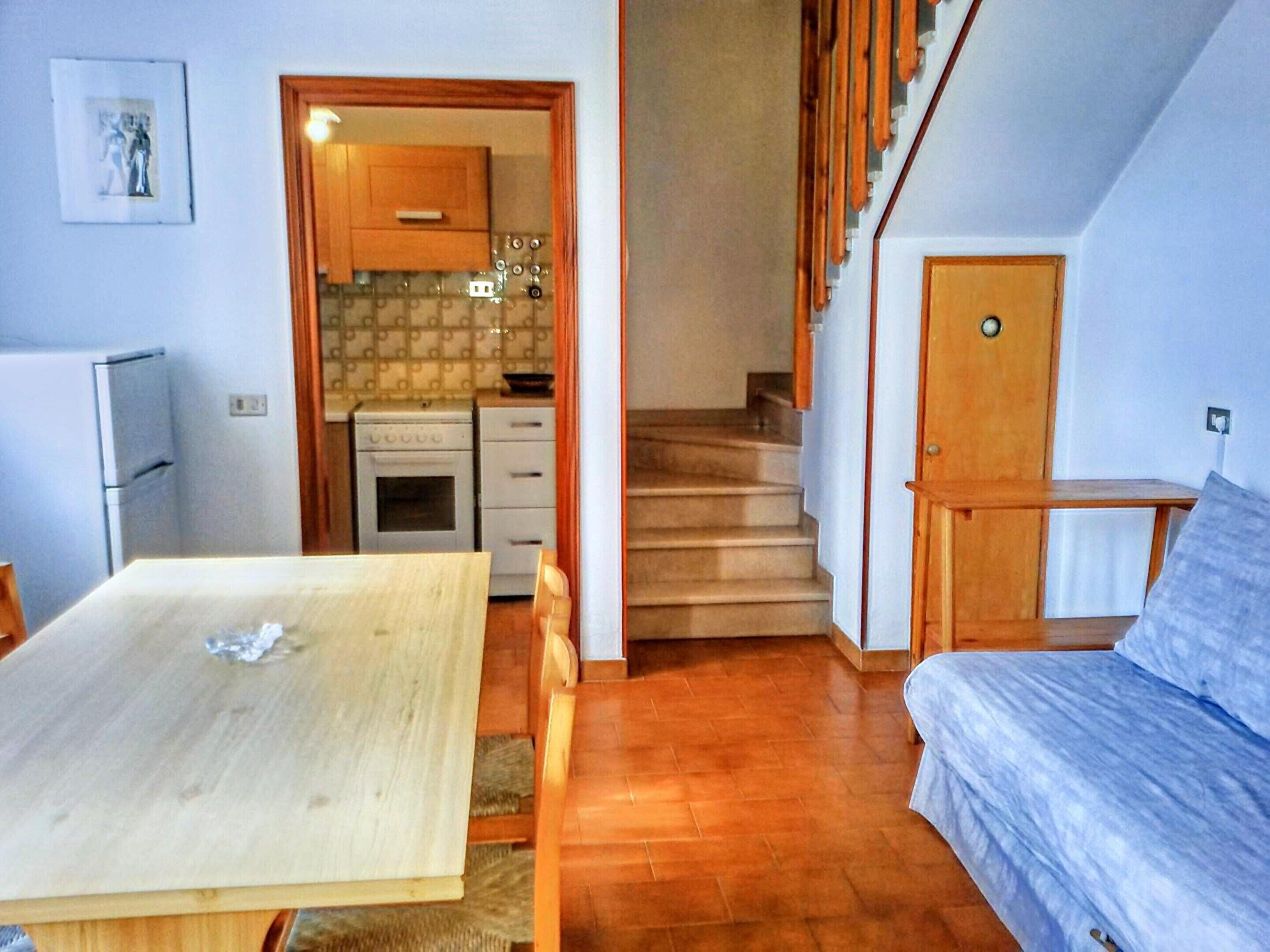 Un ambiente compatto con un tavolo da pranzo in legno, un divano, un angolo cottura dotato di fornello e pensili, e una scala.