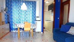 A dining area with a table and chairs, a kitchen with cabinets and appliances, and a blue sofa. - Apartment in Caorle Near Seabeach (Caorle)