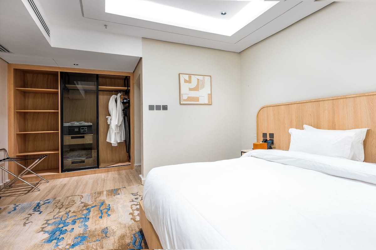 A hotel room with a skylight, a bed with white linens, a wooden headboard, a closet with a robe, and a patterned rug.