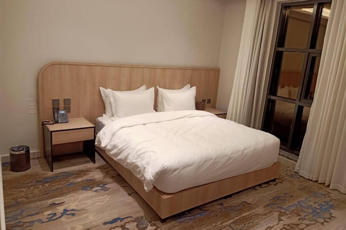 A hotel room with a wooden headboard, a bed with white bedding, a bedside table with a phone, and a window with curtains.