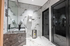 A modern bathroom with marble walls, a hand dryer, a trash bin, and a glass shower enclosure.