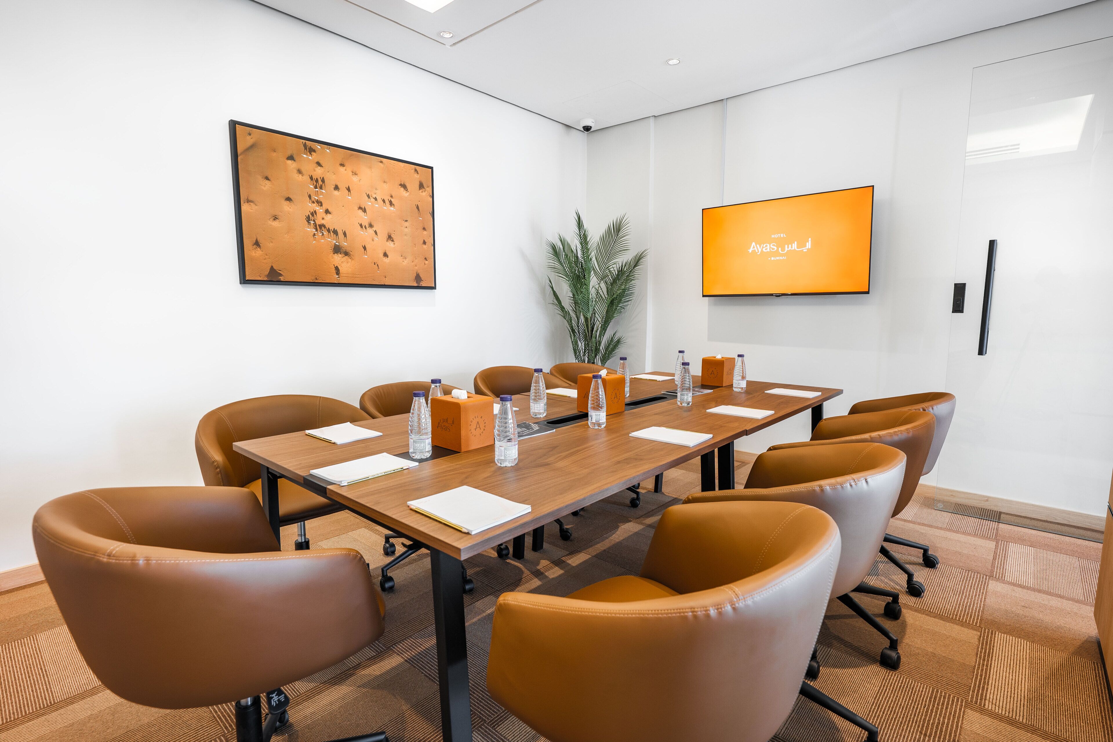 A conference room with a long table, brown leather chairs, water bottles, and a TV screen displaying text.