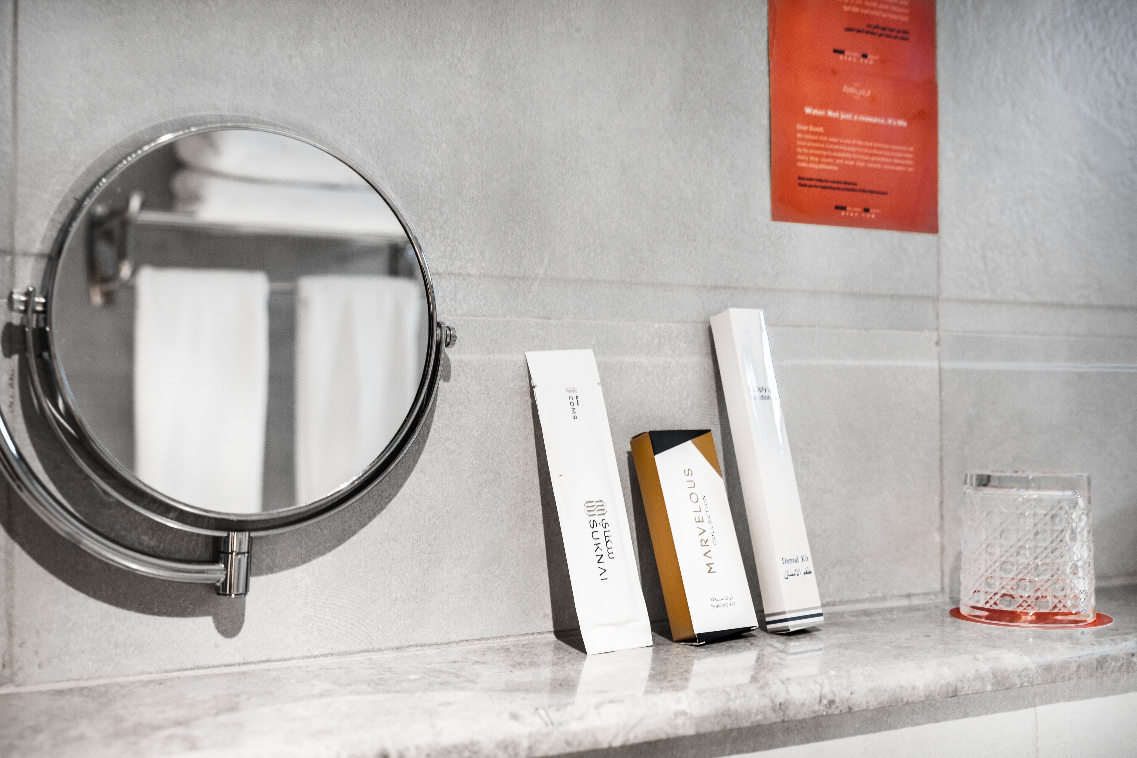 A bathroom with a round mirror, two boxes labeled 'SUKINAMI' and 'MARVELOUS,' and a glass on the countertop.