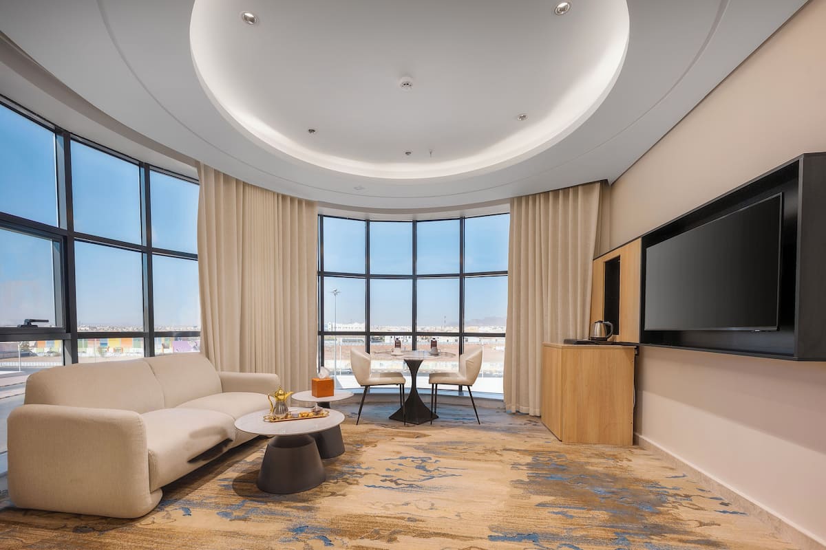 A modern hotel room with a large window, a sofa, a coffee table, a TV, and a view of the city.